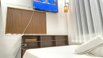 A hotel room with a bed, a TV mounted on the wall, a window with blinds, a shelf with a basket, and a wall-mounted lamp.