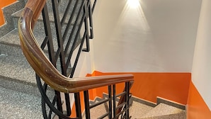 A spiral staircase with black metal railings and a wooden handrail, leading to a landing with a white wall and an orange accent wall. - Luxhome (Ho Chi Minh City)