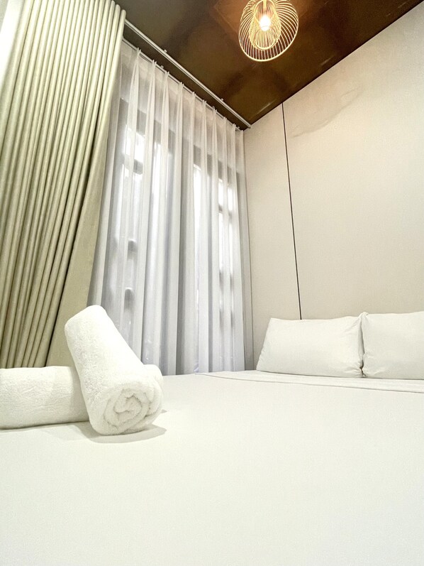 Habitación de hotel con una cama, cortinas blancas, una toalla blanca enrollada y una lámpara de techo moderna.