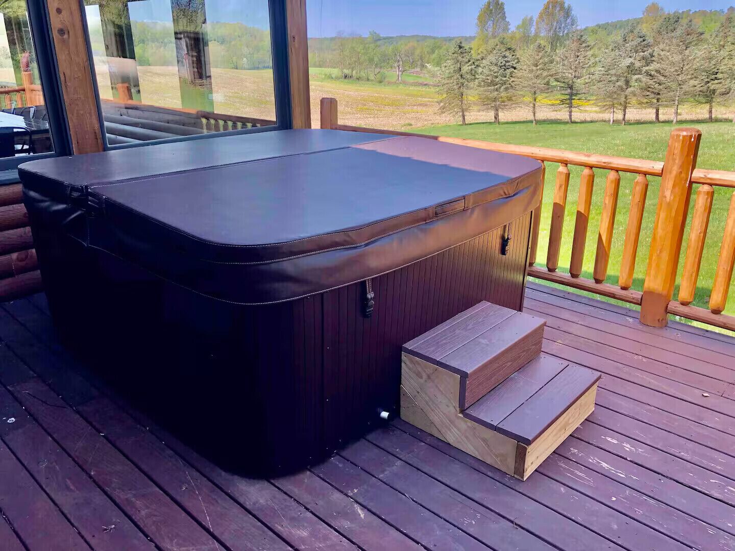 A hot tub on a wooden deck with a view of a grassy field and trees.