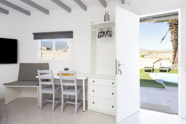 Quarto com escrivaninha branca, duas cadeiras, mesinha, televisão, janela com vista e porta para área de piscina externa.