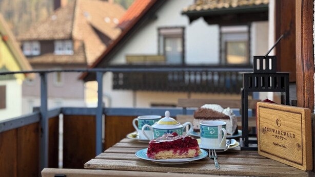 Ein Holztisch mit einem Kuchenstück, zwei Teetassen und einem Schild auf einem Balkon mit Blick auf Häuser.