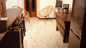 A compact kitchen with a dining table, two wicker chairs, a coffee maker, and a refrigerator.