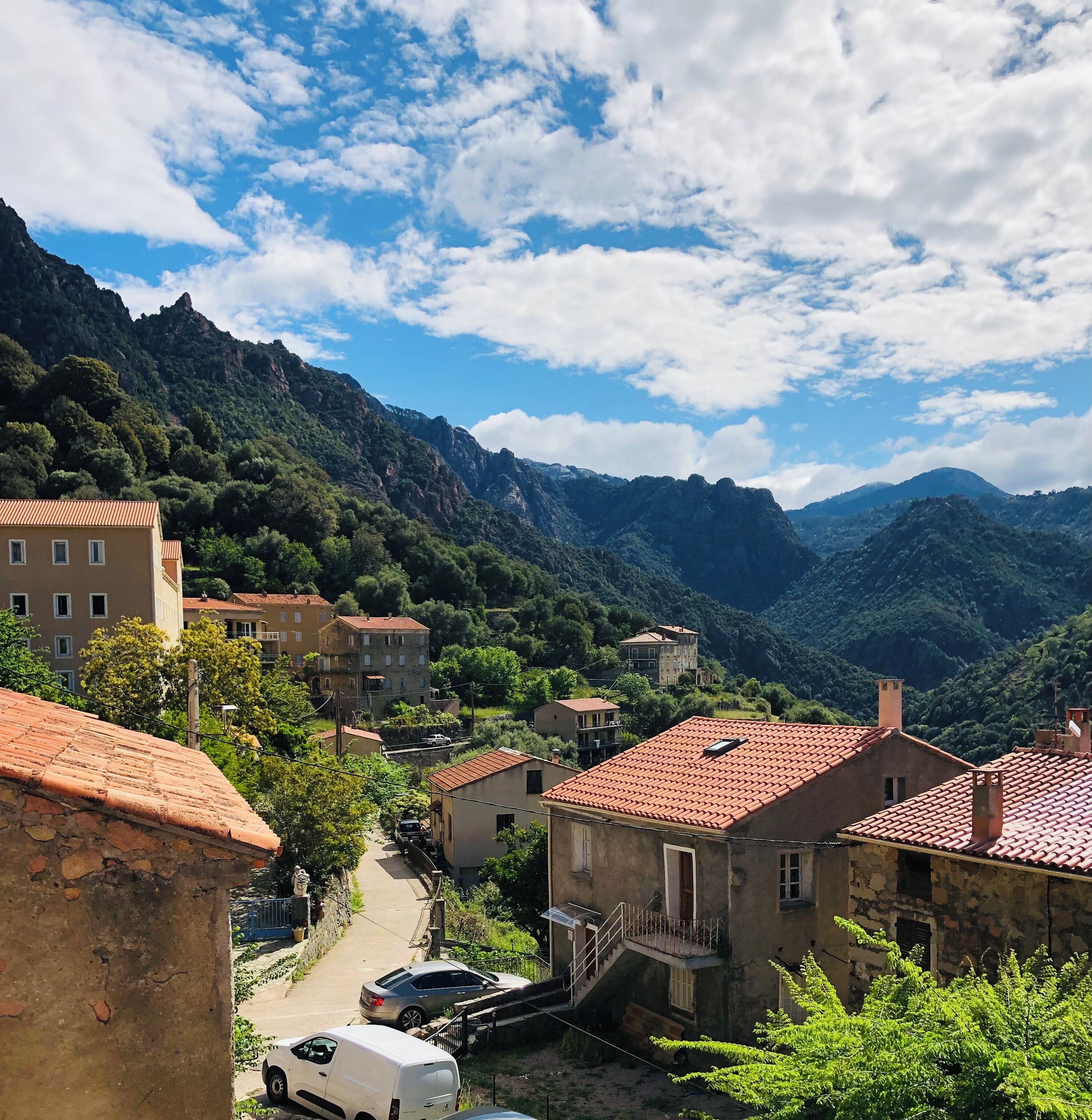 Un village perché sur une colline, avec des bâtiments aux toits de tuiles rouges, une rue étroite et, en arrière-plan, de luxuriantes montagnes verdoyantes.