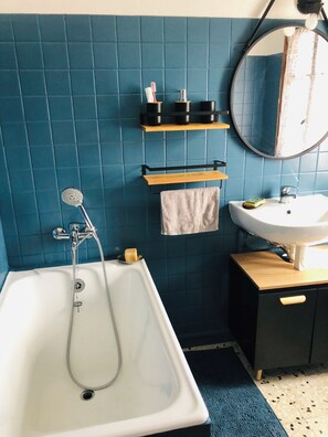 A bathroom with a bathtub, a sink, and a mirror. There are shelves with toiletries and a towel hanging on the wall. - Haven of peace (Ota)