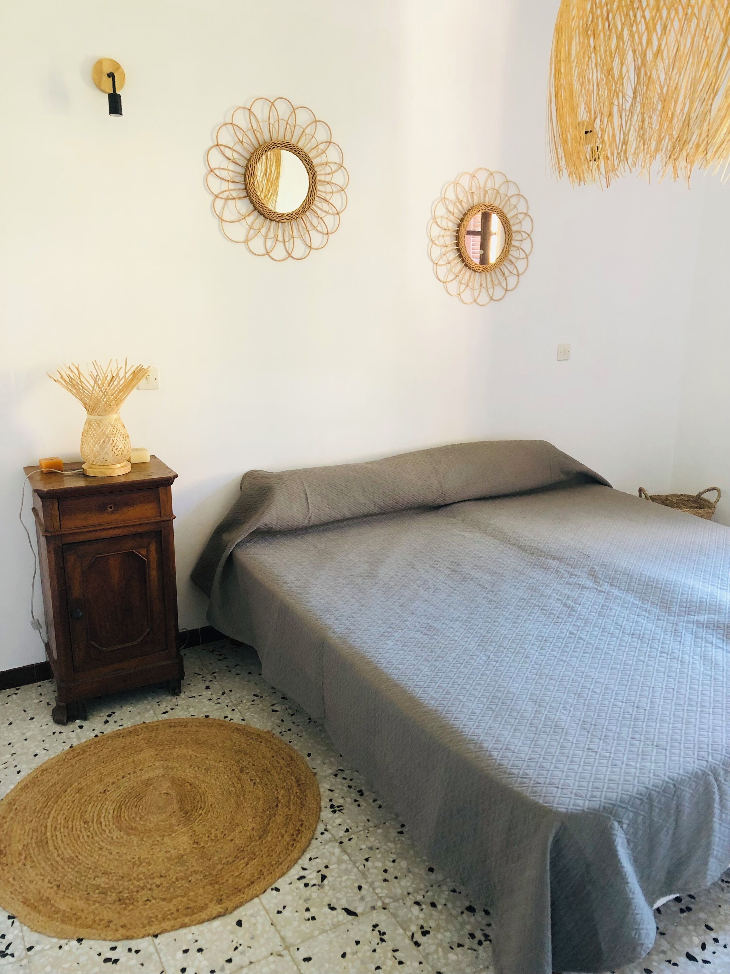Ein Schlafzimmer mit einem Bett, einem kleinen Nachttisch aus Holz, einer gewebten Fußmatte und zwei an der Wand befestigten Spiegeln.