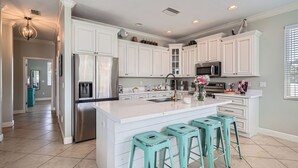 A modern kitchen with white cabinetry, a stainless steel refrigerator, a built-in microwave, and a large island with bar stools. - Location! Family Friendly, Pool, Elevator, Close to Beach (Holmes Beach)