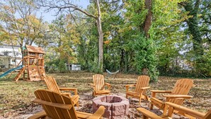 A wooden deck with Adirondack chairs arranged around a central fire pit, surrounded by trees and a playground structure.