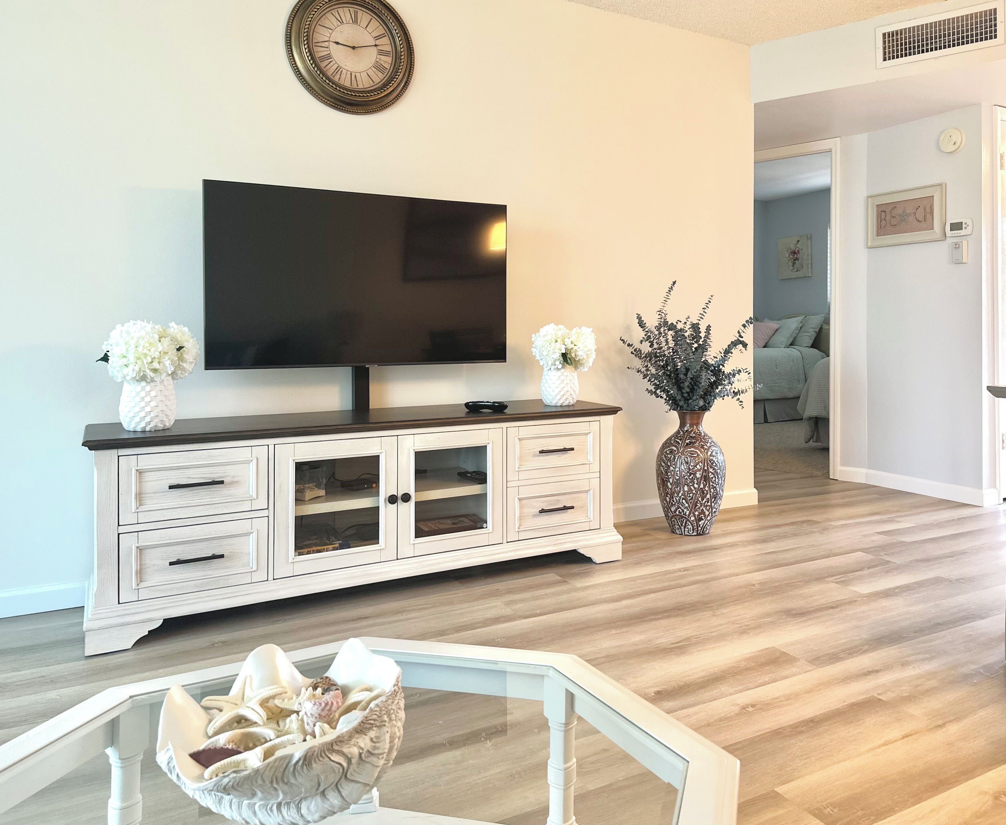 A living room with a flat-screen TV, a white cabinet with glass doors, a decorative vase, and a wicker chair with a basket.