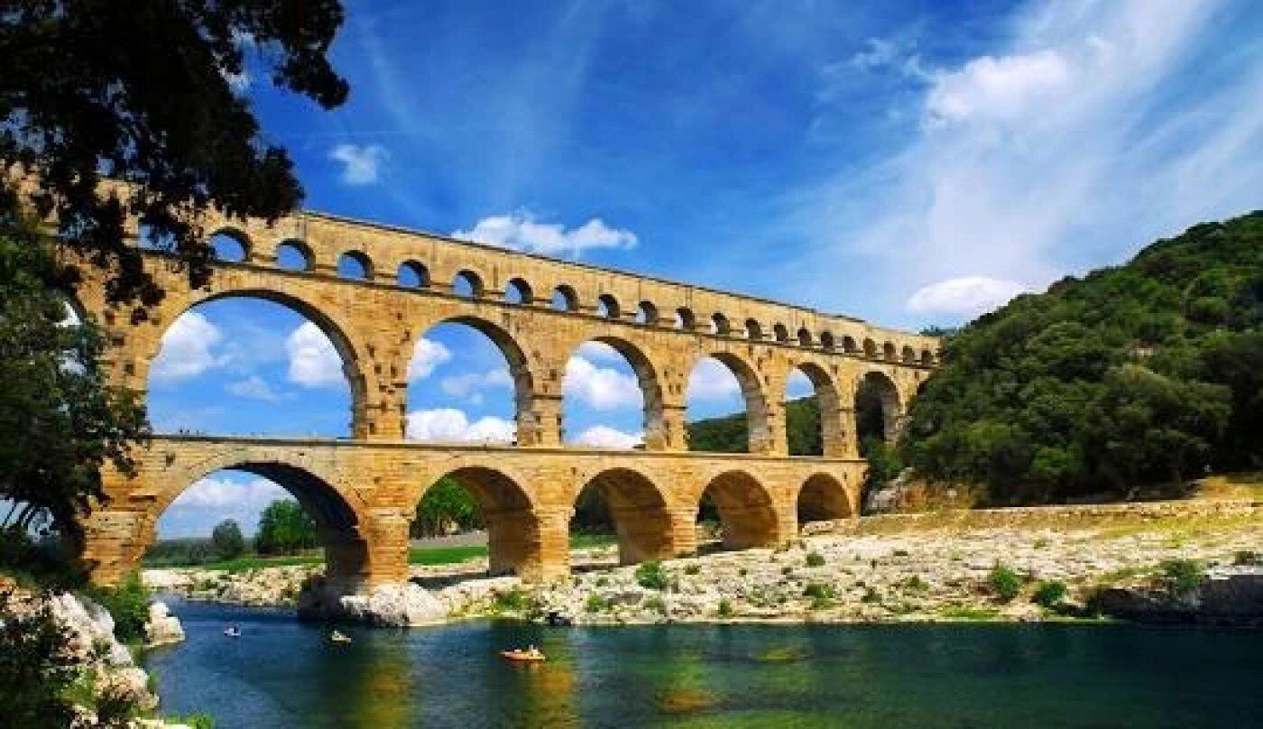 A stone aqueduct bridge over a river with clear water, surrounded by greenery and trees.