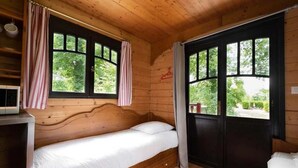 Uma cama de casal numa cabana com cabeceira de madeira, uma pequena mesa com micro-ondas e uma janela com cortinas.