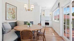 A dining area with a round wooden table, wicker chairs, a bench with cushions, a chandelier, and a kitchen in the background.