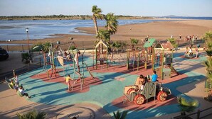 Ein Spielplatz mit verschiedenen Spielgeräten, einer kleinen Eisenbahnfahrt und einem Strand im Hintergrund.