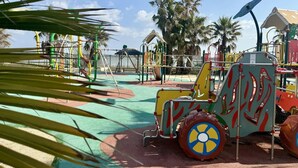 Ein Spielplatz mit verschiedenen Spielgeräten, darunter eine große Spielzone im Fahrzeugdesign, umgeben von Palmen und blauem Himmel.
