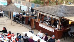 Un espace bar en plein air doté d’un toit de chaume, d’un comptoir avec un menu et de tables où sont assis les clients.