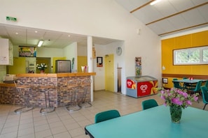 Un restaurant avec un comptoir en briques, des tabourets de bar, un réfrigérateur et un congélateur. On y trouve des chaises vertes et une table ornée d’un vase de fleurs.