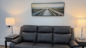 Ein modernes Wohnzimmer mit einem dunkelbraunen Ledersofa, zwei Beistelltischen mit Lampen und einem großen gerahmten Bild von einem Steg an der Wand.