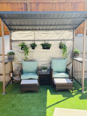 Un espace extérieur aménagé, abrité et doté de deux fauteuils en osier et d’une petite table, entouré de gazon synthétique et de plantes en pot.