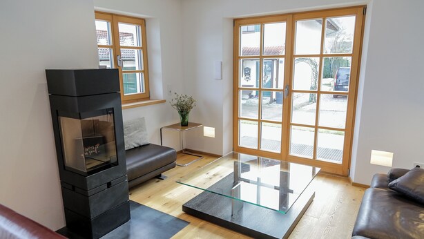 A modern living room with a black fireplace, a glass coffee table, a leather sofa, and large windows offering a view of the street. - Exclusive vacation apartment in Chiemgau (Obing)