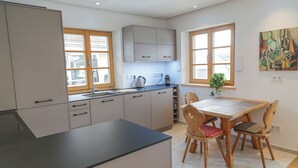 A modern kitchen with grey cabinets, a black countertop, and wooden chairs. There is a window with a view of the outside, and a painting on the wall. - Exclusive vacation apartment in Chiemgau (Obing)