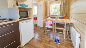 A compact kitchen with a microwave, stove, and a small refrigerator. There is a dining table with a chair and a window with curtains. - ZEcamping 3 étoiles pataugeoire mobil-home 24m2 (Pezuls)