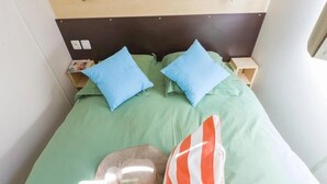 A bed with green bedding, two blue pillows, a striped pillow, a hat, and a small nightstand with a lamp. - ZEcamping 3 étoiles pataugeoire mobil-home 24m2 (Pezuls)