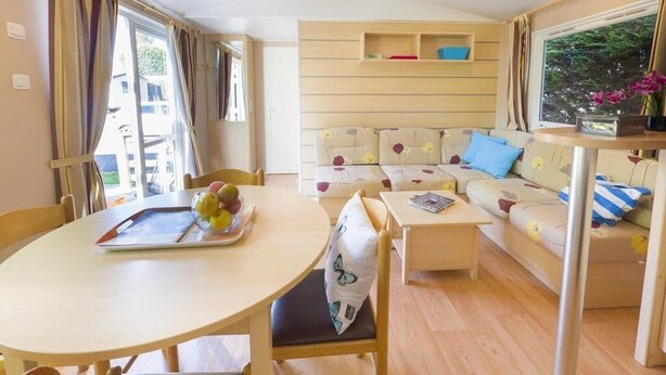 A cozy living area with a sofa, a dining table with a fruit tray, and a small coffee table. - ZEcamping 3* pataugeoire piscine locatif TV 31m2 5 pers (Pezuls)