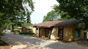 A row of rustic, single-story buildings with tiled roofs, surrounded by trees and a paved road. - ZEcamping 3* pataugeoire piscine locatif TV 31m2 5 pers (Pezuls)