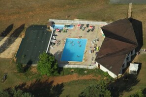 An aerial view of a building with a swimming pool surrounded by a fenced area, a tennis court, and a covered seating area.