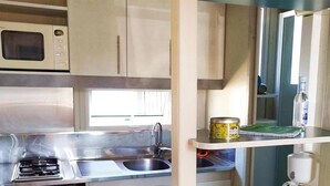 Une petite cuisine équipée d’un four à micro-ondes, d’une cuisinière, d’un évier et d’un petit réfrigérateur. On y trouve une étagère avec une conserve et une bouteille, ainsi qu’un plan de travail avec un couteau et des tomates.