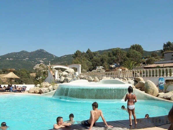 A pool area with a waterfall feature, surrounded by mountains and trees. - ZEcamping 3 étoiles à 1km de la plage piscine jacuzzi animaux acceptés (Viggianello)
