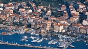Une vue aérienne d’une ville côtière, avec un port de plaisance rempli de bateaux, entourée de bâtiments résidentiels aux toits recouverts de tuiles rouges.