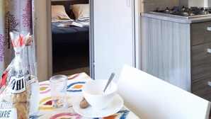 A compact hotel room with a kitchenette, a bed with a dark blue bedspread, and a table with a white bowl and a bottle of water. - ZEcamping 3* 1km plage piscine chiens acceptés locatif TV clim 25m2 (Viggianello)
