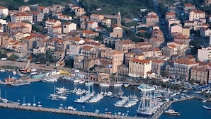 Vista aérea de un pueblo costero con un puerto deportivo lleno de barcos, rodeado de edificios residenciales con tejados de tejas rojas.