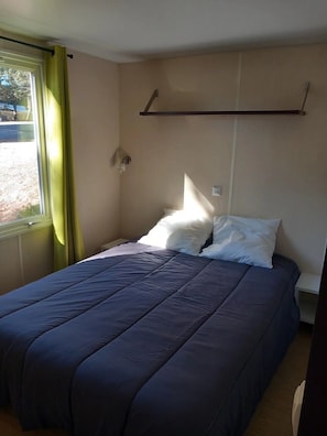 A single bed with a blue bedspread and white pillows in a room with beige walls and a window with green curtains. - ZEcamping 3 étoiles familial toboggans locatif TV BBQ clim 28m2 4 pers (Fabrezan)