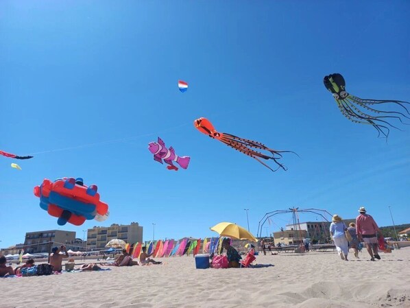A beach with people, colorful kites, and buildings in the background.