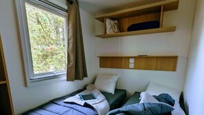 A small, compact room with two single beds, a window with curtains, and built-in shelves above the beds.