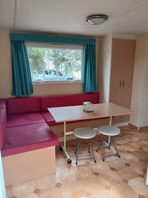 Een compacte kamer met een rode bank, een klein tafeltje, twee krukjes, een raam met gordijnen en een betegelde vloer.