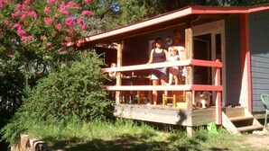 A small wooden structure with a porch, surrounded by greenery and pink flowers.