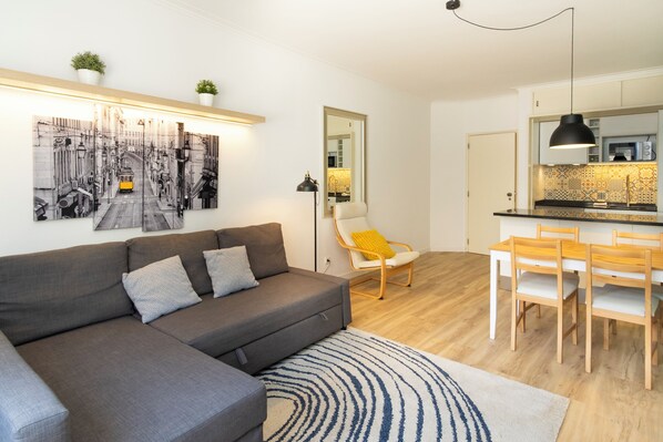 A modern living room with a grey sofa, a dining table with wooden chairs, and a kitchen area with a large mirror and pendant lighting.