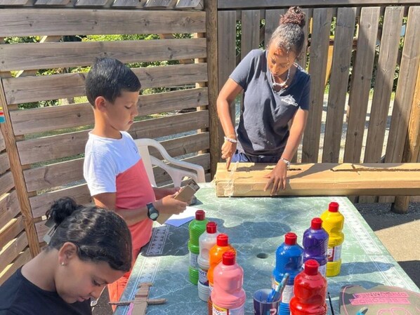 Un grupo de personas sentadas en una mesa al aire libre con botellas de distintos colores, probablemente para pintar o hacer manualidades.