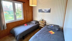 Una habitación con dos camas individuales, techo de madera, ventana con vistas al exterior y una estantería de pared con una botella y un libro.