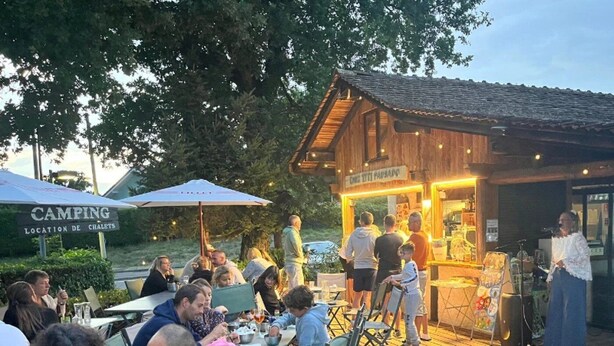 Un espace extérieur aménagé pour les repas, avec des tables et des chaises en bois, une petite scène équipée d’un microphone et un panneau indiquant l’emplacement d’un terrain de camping.
