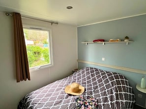 A bedroom with a bed, a window with curtains, a bedside table with a bottle, and a wall-mounted shelf with books and a plant.