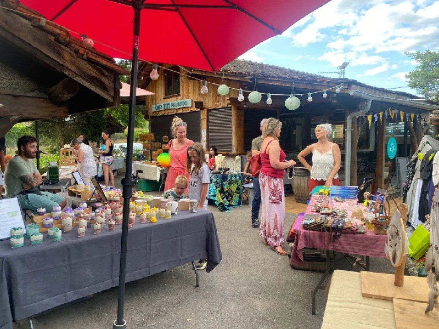 An outdoor market with various stalls selling goods, under a large red umbrella.