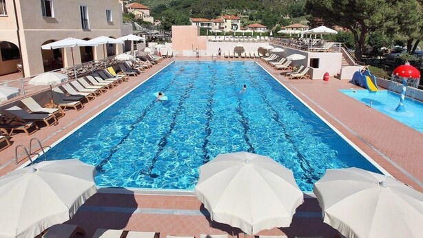 En swimmingpool med liggestole og parasoller, omgivet af bygninger og en skråning.