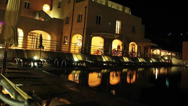 Un hôtel doté d’un espace piscine éclairé la nuit, avec des chaises longues et des reflets dans l’eau.