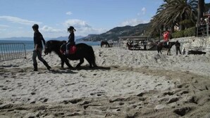 Une plage où l’on voit des gens à cheval, une zone sablonneuse et, en arrière-plan, une vue sur la mer et les collines.
