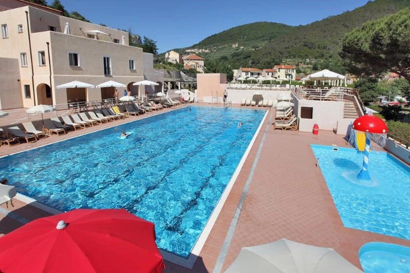 Una piscina con lettini e ombrelloni, circondata da edifici e con montagne sullo sfondo.