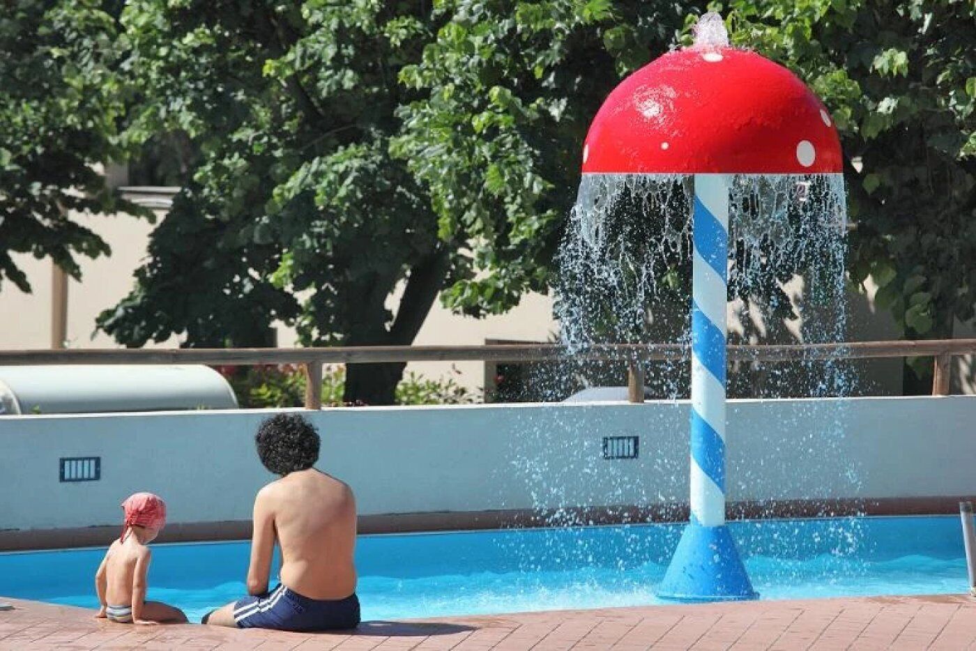 Une fontaine avec un sommet rouge et un poteau bleu, une piscine, et un enfant et un adulte assis au bord de la piscine.